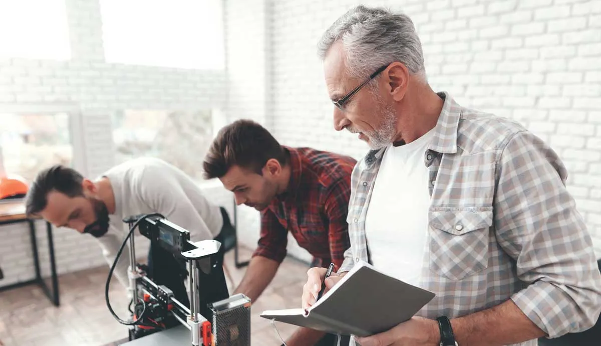 Three men work together in an office; two examine a 3D printer while the third writes in a notebook.