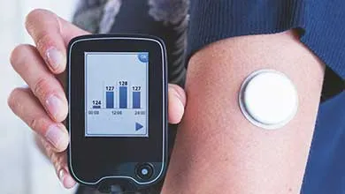 A person holds a continuous glucose monitor reader near a white, circular sensor on their upper arm. The device screen displays a blood glucose bar graph.