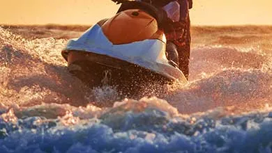 A person rides a jet ski across the water, creating splashes during a golden sunset.
