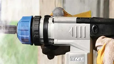Close-up of a white and black TNT rotary hammer drill being operated by a hand wearing a yellow work glove.