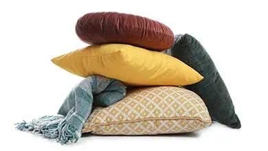 A stack of decorative pillows in brown, yellow, and green, with a teal plaid blanket, set against a white background.