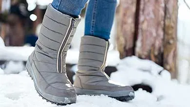 Close-up of a person standing in snow, wearing blue jeans and gray puffy winter boots with side zippers.
