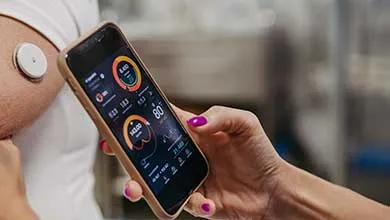 A close-up of a person’s upper arm wearing a small circular wearable sensor, while another person holds a smartphone displaying health metrics; hands adjust the sensor in a clinical or medical setting with equipment blurred in the background.