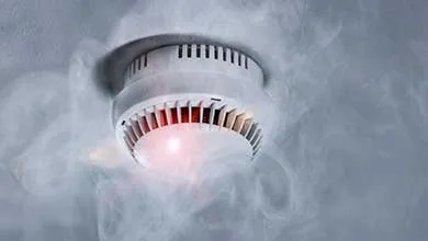 A white smoke detector with a red light on, mounted on a ceiling and surrounded by smoke.
