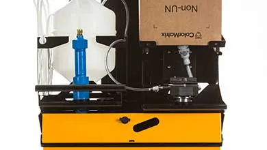 Close-up of industrial machinery with a yellow base and black frame, featuring a white fluid container and blue filter beside a cardboard box labeled "ColorMatrix" and "Non-UN".
