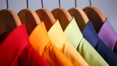 Colored shirts in red, orange, yellow, green, blue, and purple hanging on wooden hangers in a closet.