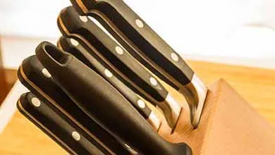 Several kitchen knives with black handles and silver rivets are stored upright in a light wood knife block, with one handle prominently in the foreground.