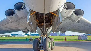 Low-angle view beneath a large four-engine jet showing four exhaust nozzles, extended multi-wheel landing gear, and open undercarriage panels revealing internal mechanical parts, with airport tarmac and grassy field in the background.