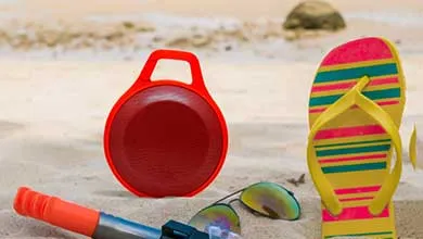 Beach items on sand: a red round container with handle, a yellow flip-flop with pink and green stripes, reflective sunglasses, and an orange and blue beach toy.